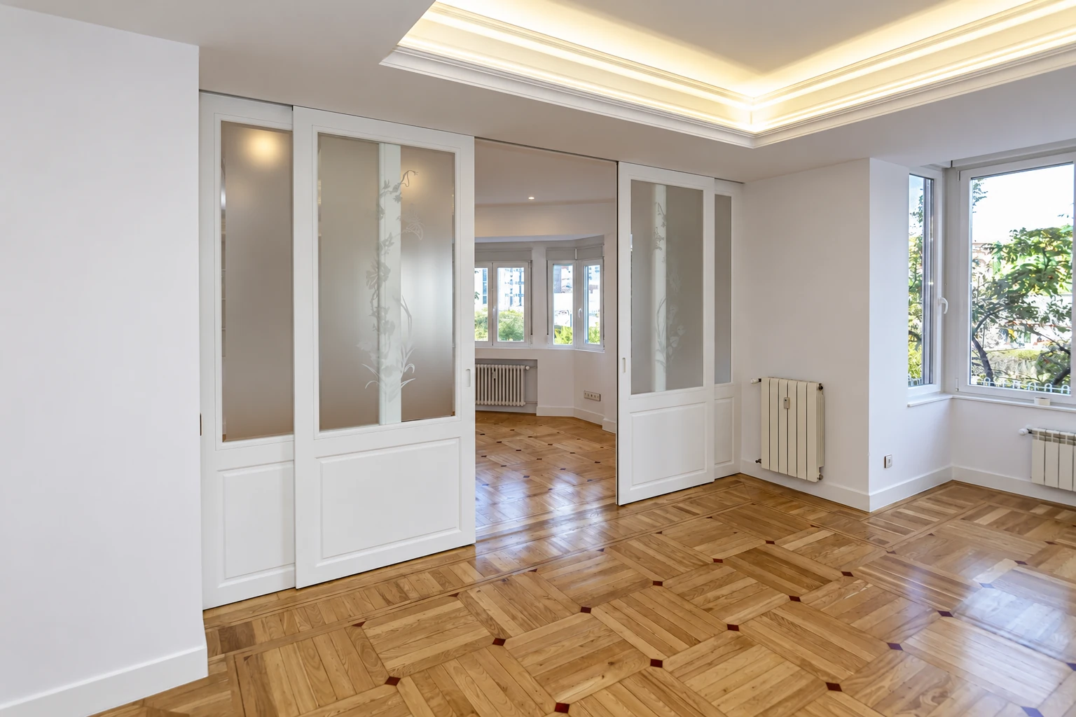 Salón con puertas correderas de vidrio decorado, parquet e iluminación indirecta en calle Galileo.