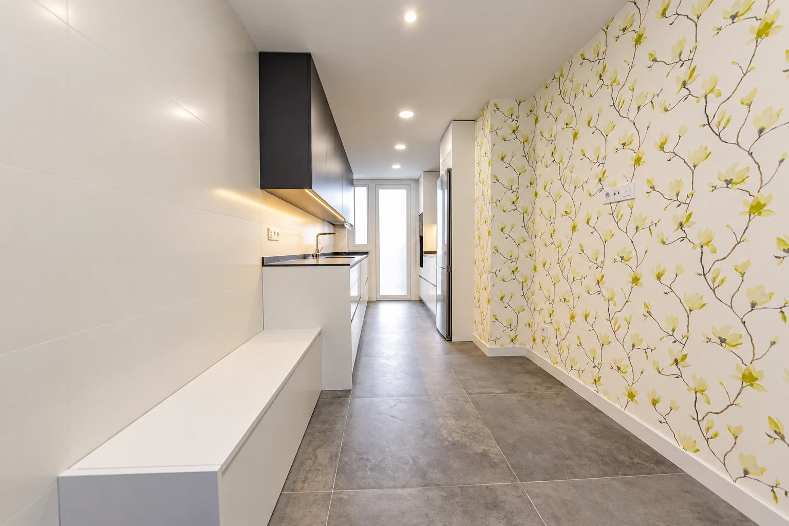 Cocina de distribución alargada con muebles blancos y papel pintado de ramas en Pozuelo de Alarcón.